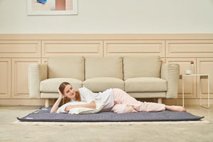 Person lying on a grey quilted floor mat in front of a beige sofa