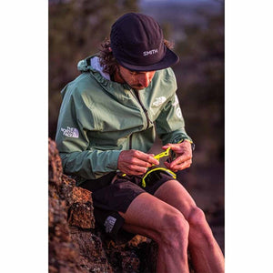 Person wearing a Smith Optics cap and green jacket sitting on a rock while holding yellow sunglasses
