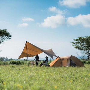 Two people sitting under a tan tarp next to a brown tent in a grassy field
