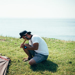 Person using a Snow Peak hammer to secure a tent stake in the grass near the ocean