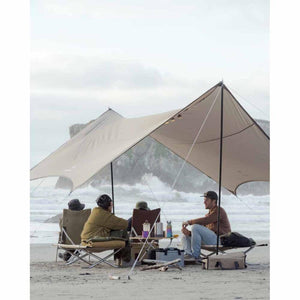 People sitting in camping chairs under a large tarp on a beach with ocean waves in the background