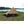 Snow Peak tan camping tarp set up over a sleeping bag and small table in a grassy field by a lake