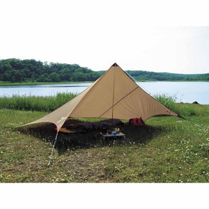 Snow Peak tan camping tarp set up over a sleeping bag and small table in a grassy field by a lake