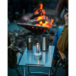 Snow Peak metal table with a canister lantern, a metal mug, and a pitcher near a campfire