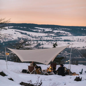 People sitting under a Snow Peak tarp by a campfire in a snowy landscape