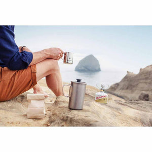 Person holding a Snow Peak mug with a camping stove and coffee bag on a rocky beach
