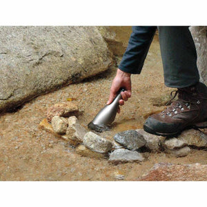 Person filling a metal water bottle in a shallow rocky stream