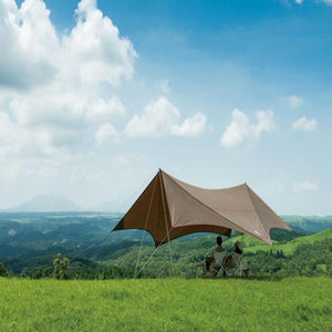Two people sitting under a brown tarp on a grassy hill overlooking a mountain landscape