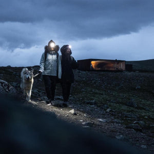 Two people wearing headlamps walking a dog on a rocky trail at dusk near a modern cabin