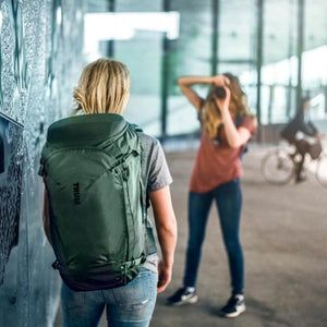 Person wearing a green Thule backpack while standing near a wall
