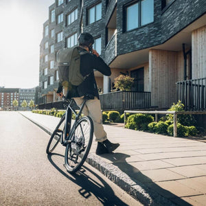 Person wearing a Thule backpack and helmet standing with a bicycle on a city sidewalk