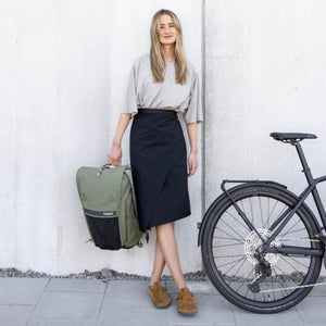 Woman standing next to a bicycle holding an olive green Thule backpack