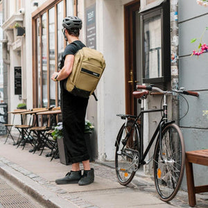 Person wearing a helmet and a tan Thule backpack standing next to a bicycle on a city sidewalk