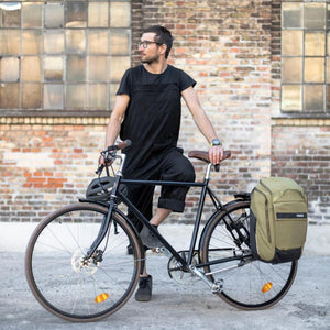 Man standing with a bicycle featuring a green Thule pannier bag against a brick wall
