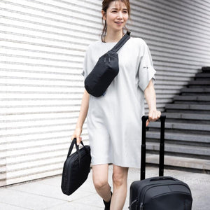 Woman wearing a black Thule sling bag while carrying a laptop sleeve and rolling luggage