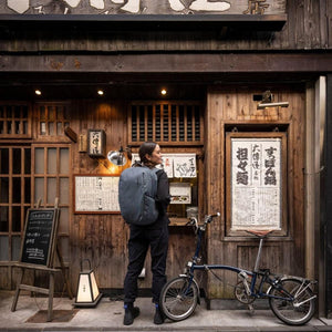 Person wearing a Thule backpack standing next to a folding bicycle in front of a wooden storefront