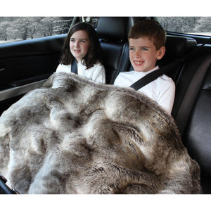 Two children sitting in a car backseat covered by a faux fur throw blanket