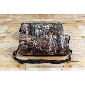 Camo hand warmer muff with Saskatchewan Original branding and black adjustable strap on wooden surface