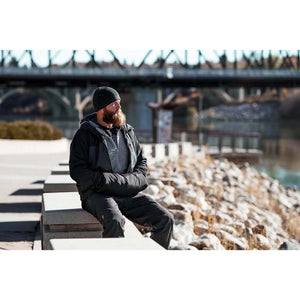 Man with beard wearing black beanie and jacket sitting on concrete wall by river