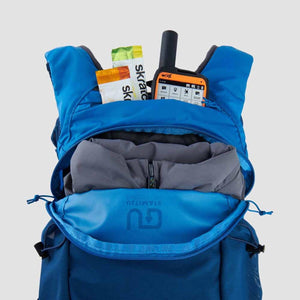 Blue backpack with open main compartment containing a grey jacket Skratch Labs snack and a satellite communicator