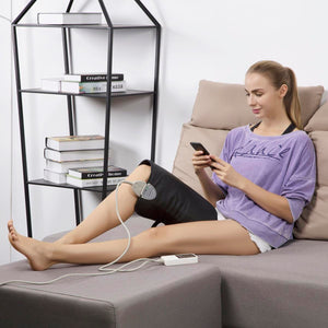 Woman sitting on a sofa using a black electric knee heating pad while looking at a smartphone