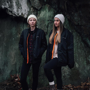 Two women wearing Venture Heat black bomber jackets and beanies standing in front of a rock wall