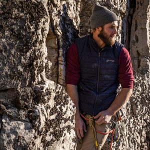 Man wearing a Venture Heat black vest and beanie leaning against a rock face with climbing gear