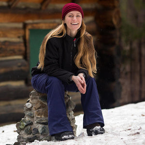 Woman with long hair wearing a red beanie and black jacket sitting on a stone pillar in the snow