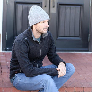 Man wearing a grey cable knit beanie and a black zip up sweater sitting on brick steps