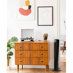 Black Vornado tower fan standing on a wooden floor next to a mid-century modern dresser