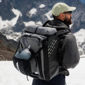 Person wearing a grey Kulkea backpack with a helmet stored in the exterior pocket against a snowy mountain background