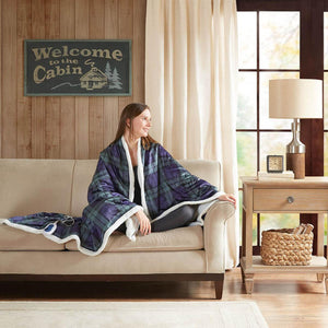 Woman sitting on a sofa draped in a Woolrich green and blue plaid heated throw blanket with white sherpa lining